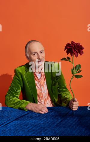 Positiver und modischer Senior-Mann in grünem Samtblazer mit roter Pfingstrose, sitzt mit blauem Velours am Tisch und lächelt vor der Kamera Stockfoto