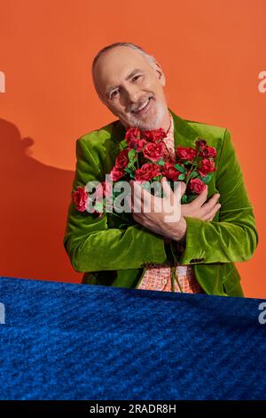 Ein überfreuliches männliches Senior-Model in einem grünen Samtblazer, der einen Strauß roter Rosen umarmt und in der Nähe des Tisches vor der Kamera lächelt, mit blauem Velourtuch auf leuchtendem o Stockfoto