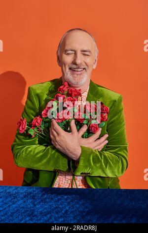 Ein überfreulicher Senior-Mann in grünem Samtblazer umarmt rote Rosen und lächelt mit geschlossenen Augen am Tisch mit blauem Velours-Tuch auf leuchtend orangefarbenem Backgro Stockfoto