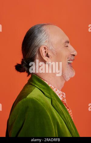 Seitenansicht eines aufgeregten und modischen älteren Modells, das lacht, mit geschlossenen Augen auf leuchtend orangefarbenem Hintergrund, Porträt eines fröhlichen und bärtigen Seniorenmanns i. Stockfoto