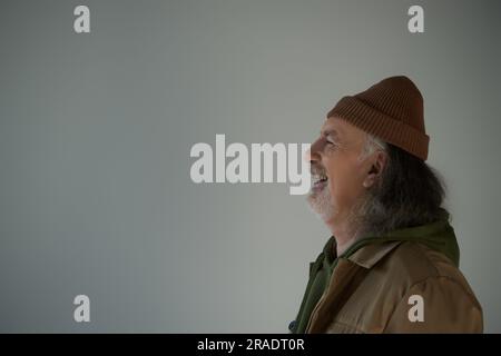 Seitenansicht eines aufgeregten, grauhaarigen und bärtigen Seniorenmannes mit Beanie-Hut und brauner Jacke, der auf grauem Hintergrund lacht, fröhliche und modische alternde Conce Stockfoto