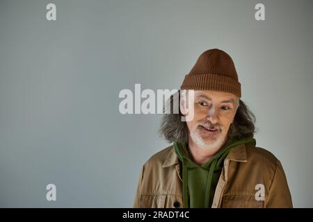 Grauhaariger und bärtiger Senior-Mann, aufmerksam und lächelnd, mit Beanie-Hut und brauner Jacke auf grauem Hintergrund, Hipster-Mode, fröhlich und modisch Stockfoto