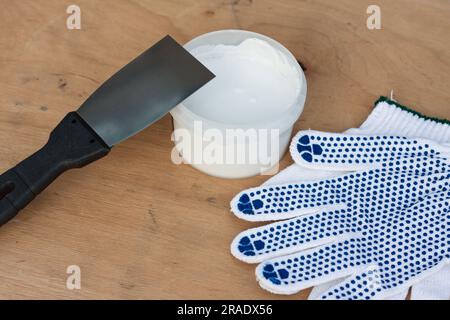 Baugruppensatz, Bauspachtel, Dose mit weißem Kitt, Schutzhandschuhe, auf Sperrholzoberfläche. Stillleben. Einbausatz für Kitt. Stockfoto