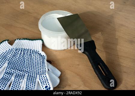 Baugruppensatz, Bauspachtel, Dose mit weißem Kitt, Schutzhandschuhe, auf Sperrholzoberfläche. Stillleben. Einbausatz für Kitt. Stockfoto