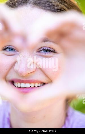 Erleben Sie die fesselnde Schönheit und strahlende Freude, während eine junge Frau mit ihren Händen ein Herzsymbol kreiert und mit einem reinen Zauber hindurchblickt. Dieses Porträt fängt sie in einem Moment der aufrichtigen Verbindung ein, in dem Liebe und Freude in ihr aufkommen. Das Herzsymbol repräsentiert ihren offenen und mitfühlenden Geist und symbolisiert die Tiefe ihrer Emotionen. Ihr fesselnder Blick spiegelt die wahre Schönheit wider, die sich aus Liebe und Freude ergibt. Erleben Sie die Kraft ihres Ausdrucks, während sie einen visuellen Rahmen der Zuneigung erzeugt, der Sie einlädt, die Wärme und die positive Einstellung zu spüren, die von ihr ausgeht. - Nein Stockfoto