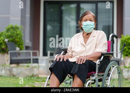 Seniorin trägt medizinische Maske für Protect Coronavirus (Covid-19), die im Rollstuhl draußen im Haus sitzt Stockfoto