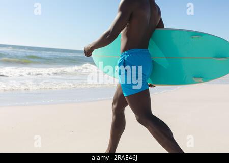 Mittlerer Abschnitt eines fitten afroamerikanischen Mannes mit Surfbrett auf dem Weg vom sonnigen Strand zum Meer Stockfoto