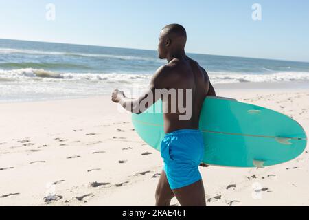 Fit-afroamerikanischer Mann mit Surfbrett auf dem Weg vom sonnigen Strand zum Meer Stockfoto