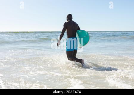 Ein afroamerikaner mit Surfbrett, der am sonnigen Strand ins Meer geht Stockfoto