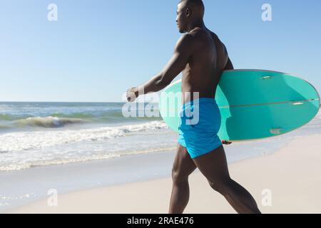 Fit-afroamerikaner mit Surfbrett, der über den sonnigen Strand zum Meer läuft, Kopierraum Stockfoto