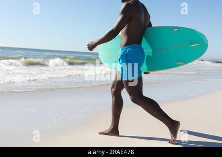 Tiefer Teil eines afroamerikaners mit Surfbrett, der über den sonnigen Strand zum Meer läuft, Kopierraum Stockfoto