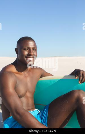 Porträt eines glücklichen afroamerikanischen Mannes, der am sonnigen Strand sitzt und ein lächelndes Surfbrett hat Stockfoto