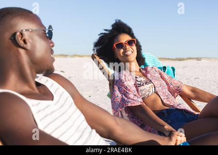 Glückliches afroamerikanisches Paar, das auf Liegestühlen sitzt und sich am sonnigen Strand die Hände hält Stockfoto