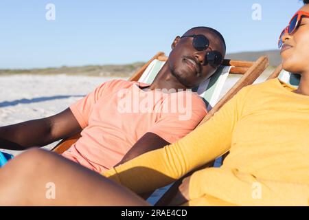 Glückliches afroamerikanisches Paar, das auf Liegestühlen sitzt und sich am sonnigen Strand entspannt Stockfoto