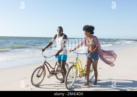 Glückliches afroamerikanisches Paar, das mit dem Fahrrad am sonnigen Strand auf dem Meer spaziert Stockfoto