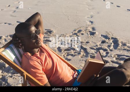 Ein glücklicher afroamerikanischer Mann mit Sonnenbrille, der im Liegestuhl sitzt und am sonnigen Strand ein Buch liest Stockfoto