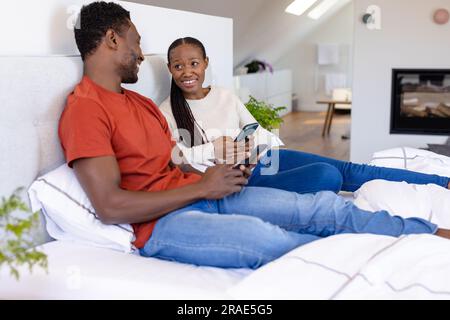 Glückliches afroamerikanisches Paar, das Smartphones benutzt, die auf dem Bett im Schlafzimmer liegen Stockfoto