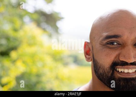 Halbes Porträt eines lächelnden, kahlen, birassischen Mannes mit Bart vor Baumwipfeln, Kopierraum Stockfoto