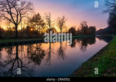 Erleben Sie die beeindruckende Schönheit des Meisterwerks der Natur, während ein dramatischer und farbenfroher Sonnenuntergang eine atemberaubende Leinwand malt, mit Baumsilhouetten, die sich im ruhigen Wasser des Flusses widerspiegeln. Der Himmel wird zu einer lebhaften Symphonie von Farbtönen, mit feurigen Orangen, tiefen Purpur- und Goldtönen, die eine fesselnde Darstellung schaffen. Die majestätischen Silhouetten der Bäume schmücken den Horizont, ihre Zweige reichen in Richtung Himmel, während der Fluss darunter ihre Eleganz spiegelt. Erleben Sie die Ruhe und Stille dieses magischen Moments, in dem die Konvergenz von Farben, Reflexionen und der Anmut der Natur entsteht Stockfoto