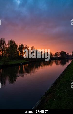 Erleben Sie die beeindruckende Schönheit des Meisterwerks der Natur, während ein dramatischer und farbenfroher Sonnenuntergang eine atemberaubende Leinwand malt, mit Baumsilhouetten, die sich im ruhigen Wasser des Flusses widerspiegeln. Der Himmel wird zu einer lebhaften Symphonie von Farbtönen, mit feurigen Orangen, tiefen Purpur- und Goldtönen, die eine fesselnde Darstellung schaffen. Die majestätischen Silhouetten der Bäume schmücken den Horizont, ihre Zweige reichen in Richtung Himmel, während der Fluss darunter ihre Eleganz spiegelt. Erleben Sie die Ruhe und Stille dieses magischen Moments, in dem die Konvergenz von Farben, Reflexionen und der Anmut der Natur entsteht Stockfoto