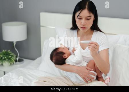 Gestresste Mutter misst die Temperatur mit dem Thermometer des kranken Babys und weint auf dem Bett Stockfoto