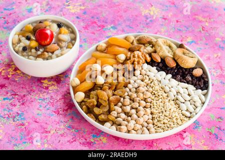 Ungekochte Ashura-Zutaten: Kichererbsen, Weizen, Reis und weiße Bohnen, Nüsse und süße Früchte auf weißem Teller auf rosafarbener Oberfläche mit einer Schüssel Noah's Stockfoto