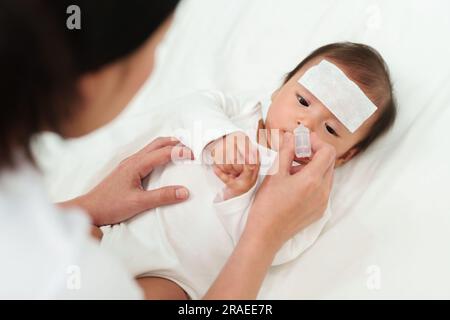 Ein krankes Baby mit kalter Fieberpolster bekommt Nasentropfen von der Mutter auf dem Bett Stockfoto