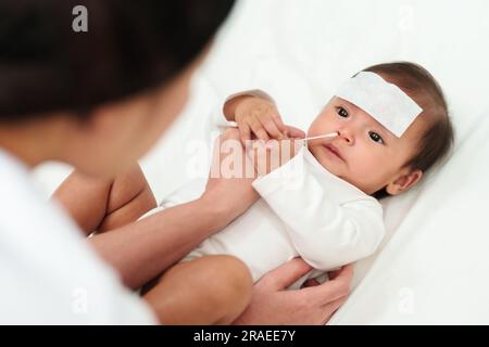 Mutter, die Wattestäbchen benutzt, um die Nase eines kranken Babys mit kaltem Fieber-jel-Pad auf einem Bett zu reinigen Stockfoto