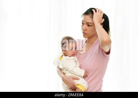 Gestresste Mutter, die mit ihrem Baby auf Fensterhintergrund hält Stockfoto