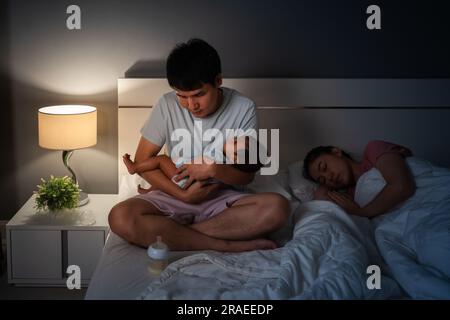 Schläfriger und müder Vater hält ein Neugeborenes im Arm, während seine Frau nachts in einem Bett schläft Stockfoto