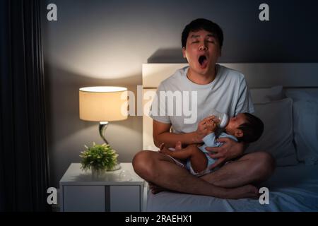 Schlafmütze Vater gähnt und füttert Milchflasche an Neugeborenes nachts auf einem Bett Stockfoto