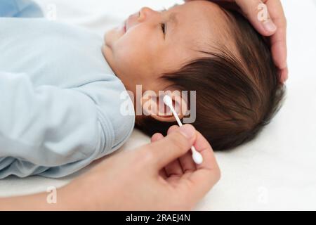 Nahaufnahme der Mutter mit Wattestäbchen, um das Ohr des Neugeborenen auf einem Bett zu putzen Stockfoto