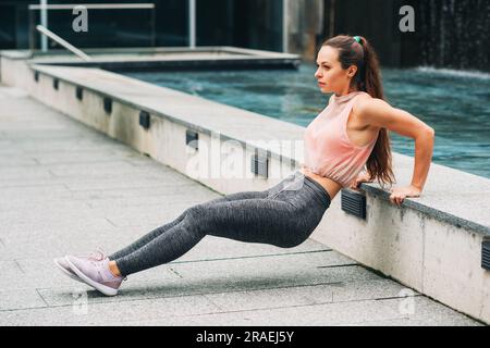 Außenporträt einer jungen, wunderschönen Frau, die Tricep Dips macht, modische Aktivkleidung trägt, Sportlermodel im urbanen Hintergrund posiert, Sport-Fashi Stockfoto