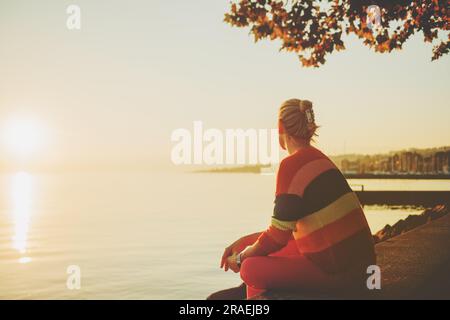 Wunderschöne Frau, die sich am See bei Sonnenuntergang ausruht, an warmen Abenden, mit farbenfrohem Pullover, Rückansicht Stockfoto