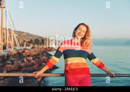 Wunderschöne Frau, die sich am See bei Sonnenuntergang ausruht, an warmen Abenden, in farbenfrohem Pullover Stockfoto