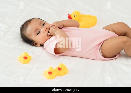 Baby, das die Hände mit den Saugfingern in den Mund steckt und ein gelbes Gummiente-Spielzeug auf einem Bett auflegt Stockfoto