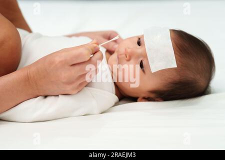 Mutter, die Wattestäbchen benutzt, um die Nase eines kranken Babys mit kaltem Fieber-jel-Pad auf einem Bett zu reinigen Stockfoto