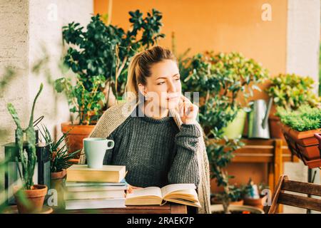 Junge schöne Frau, die sich auf dem gemütlichen Balkon entspannt, ein Buch liest, einen warmen Strickpullover trägt, Tee oder Kaffee auf einem Stapel Bücher Stockfoto