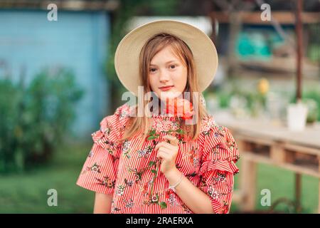 Sommerportrait eines hübschen kleinen Mädchens mit rotem Streifenkleid, großem Strohhut, Holdin-Rosenblume Stockfoto