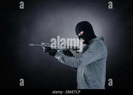 Ein Krimineller trägt schwarze Balaclava und Kapuze mit Sturmgewehr auf dunklem, nebligen Hintergrund Stockfoto