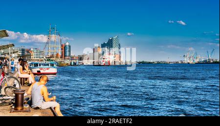 Die Menschen entspannen sich entlang der Uferpromenade am Hamburger Hafen mit der Elbphilharmonie und den Hafenkranichen im Hintergrund Stockfoto