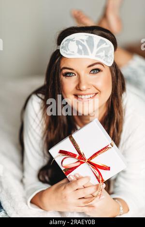 Innenporträt einer glücklichen jungen Frau, die sich im Schlafzimmer entspannt, Pyjama und Schlafmaske trägt, ein Geschenk in der Hand hat Stockfoto