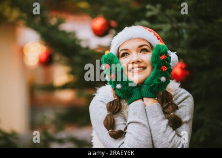 Festliches Porträt einer glücklichen jungen Frau, die draußen am Weihnachtsbaum posiert und grüne Fäustlinge trägt Stockfoto