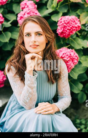 Außenporträt einer schönen Frau, die in rosa Hortensien posiert und ein blaues Kleid trägt Stockfoto