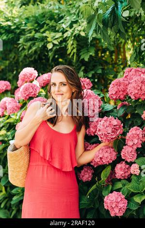 Außenporträt einer schönen Frau, die in rosa Hortensien posiert und ein rotes Kleid trägt Stockfoto