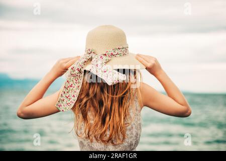 Außenporträt eines hübschen jungen Mädchens mit rotem Haar, mit großem Hut, Familienurlaub mit Kindern am Meer. Rückansicht Stockfoto