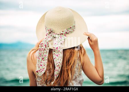 Außenporträt eines hübschen jungen Mädchens mit rotem Haar, mit großem Hut, Familienurlaub mit Kindern am Meer. Rückansicht Stockfoto