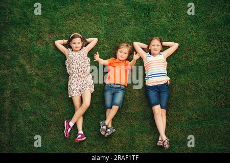 Fröhliche Kinder, die draußen Spaß haben. Kinder spielen im Sommerpark. Ein kleiner Junge und zwei Mädchen liegen auf grünem, frischem Gras Stockfoto