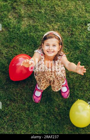 Fröhliches kleines Mädchen, das mit bunten Ballons draußen spielt, Draufsicht Stockfoto