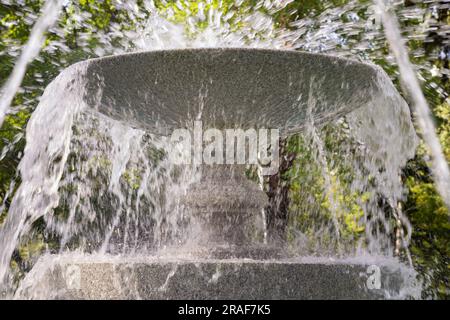 Nahaufnahme Granitschüssel im Brunnen mit Spritzwasser und Wasserstrahlen auf grünem Parkhintergrund, komfortable städtische Umgebung. Großer runder Steinfußboden Stockfoto